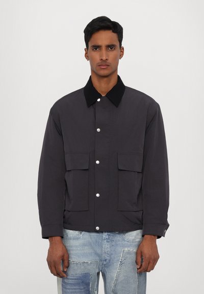 Veste gris foncé en tissu lisse, avec un col noir, une fermeture à boutons-pression, et deux poches avant avec rabats.