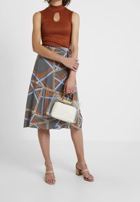 Top senza maniche a coste marrone con dettaglio a oblò, gonna midi a fantasia nei toni del nero, blu e marrone, borsa trasparente con manico in legno, tacchi a strappo beige.