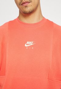Sweatshirt en tricot corail avec un col rond, des manches longues et un logo à l'avant en blanc indiquant "NIKE AIR." Tissu texturé avec des accents en panneaux latéraux.