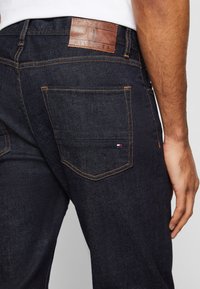 Jeans in denim blu scuro con una toppa in pelle marrone sul cinturino con la scritta "TOMMY HILFIGER" in rilievo, con cuciture dorate e due tasche posteriori.