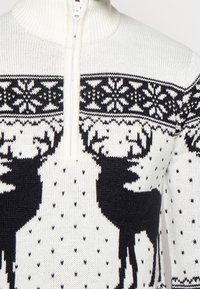 Stickad tröja i krämfärg med svarta renar och snöflinge-mönster. Har hög krage och dragkedja. Tyg med struktur och prickdetaljer.