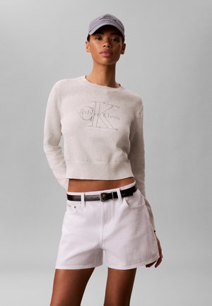 Kobieta ubrana w jasnobeżowy sweter Calvin Klein, białe szorty z czarnym paskiem i jasnoszary bejsbolowy czapka Calvin Klein.