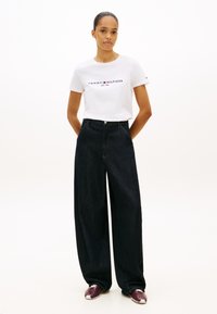 Biały bawełniany T-shirt z logo "TOMMY HILFIGER", zestawiony z ciemnoniebieskimi, szerokimi jeansami z wysokim stanem oraz bordowymi sneakersami.