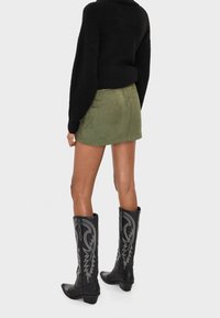 Schwarzer Strickpullover, grüner Wildleder-Minirock und schwarze Cowboy-Stiefel mit silberner Stickerei, von hinten gezeigt.