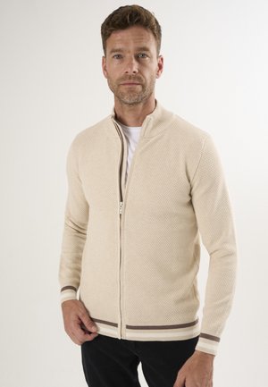Cárdigan de punto beige con cremallera y patrón texturizado, puños y dobladillo acanalados que presentan rayas marrones. Lleva puesto sobre una camiseta blanca.