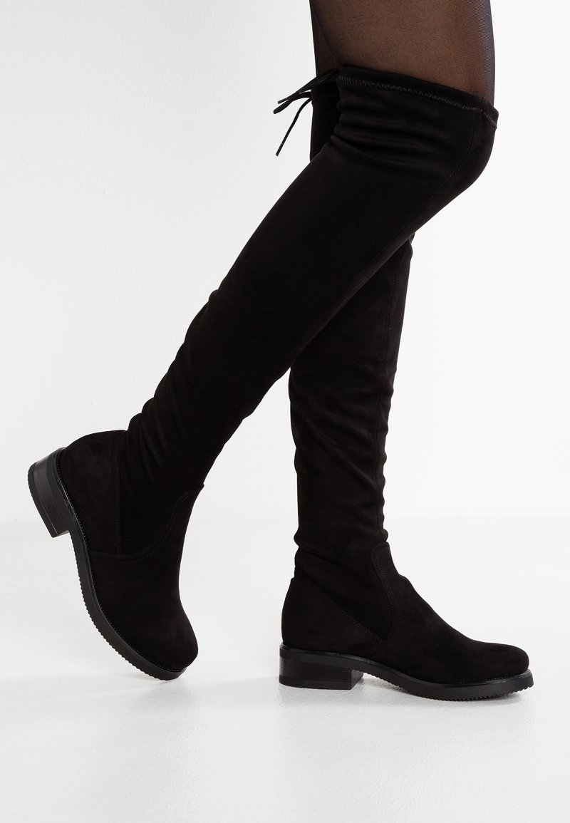 Bottes en daim noir au-dessus du genou avec des petits talons carrés portées avec des collants noirs transparents, un pied légèrement levé comme en marchant.