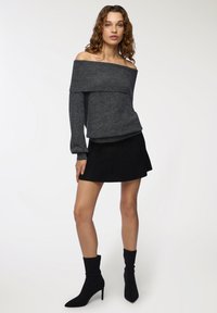 Donna con capelli ricci che indossa un maglione grigio scuro con scollatura a spalla, una mini gonna nera e stivali neri con tacco alla caviglia, in piedi davanti a uno sfondo bianco.