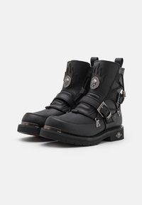 Botas de tobillo de cuero negro con herrajes plateados, hebillas y detalles de llamas, con una suela texturizada y una placa decorativa con el logo.