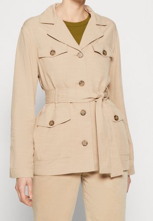 Chaqueta de utilidad beige con cintura ceñida, mangas largas, cuello de pico, dos bolsillos en el pecho y botones a juego, hecha de tela texturizada.