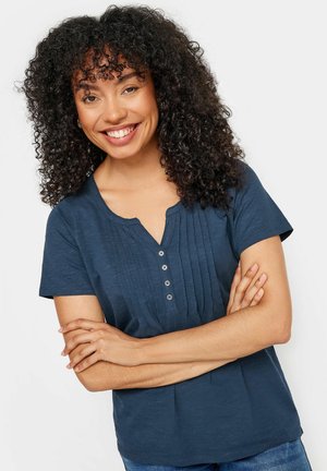 Uśmiechnięta kobieta o kręconych włosach, ubrana w granatową bluzkę z krótkim rękawem i guzikami, ze skrzyżowanymi ramionami w spokojnej pozie na jednolitym tle.