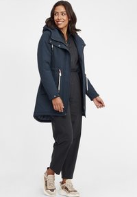 Granatowy płaszcz do długości średniej z kapturem, zamkami błyskawicznymi i sznurkiem; ma teksturowany materiał i kieszenie boczne, w połączeniu z ciemnymi spodniami i trampkami.