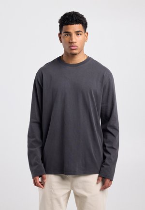 Jonge man met kort krullend haar, draagt gouden oorbellen en een ketting, een donkergrijs shirt met lange mouwen en lichtbeige broek, staand tegen een witte achtergrond.