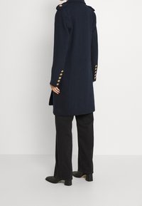 Cappotto sartoriale blu navy con spalline, dettagli in bottoni dorati e una vestibilità strutturata, abbinato a pantaloni neri svasati e stivali.