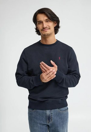 Uomo con capelli castano scuro di media lunghezza, indossa un maglione blu navy e jeans, in piedi e che tocca delicatamente la mano sinistra con la destra.