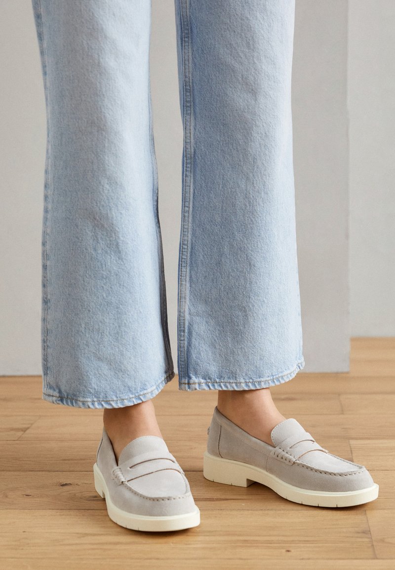Jeans de pierna ancha de color azul claro combinados con mocasines de ante beige, de pie en un suelo de madera, con un fondo de pared lisa.