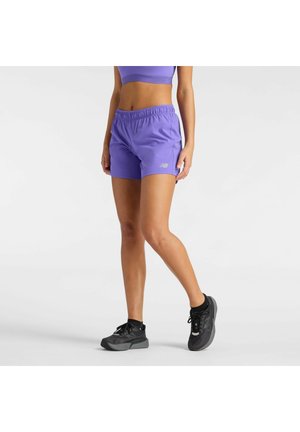 Frau trägt lila Sportshorts und Top mit schwarzen Laufschuhen, steht mit einem leicht gebeugten Bein vor weißem Hintergrund.