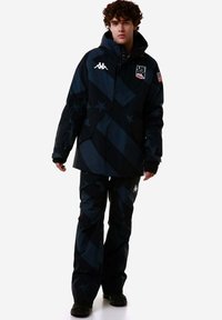 Giacca invernale nera e blu scuro con motivo a stelle, dotata di logo, abbinata a pantaloni coordinati. Il tessuto appare testurizzato e resistente.