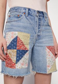 Denim shorts with patchwork design featuring floral and geometric patterns in red, yellow, blue, and white. Frayed hem detail.