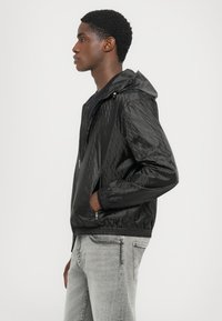 Svart lättviktsjacka med huva, med ett strukturerat mönster, dragkedjestängning och elastiska ärmslut. Bärs över en svart skjorta med grå jeans.