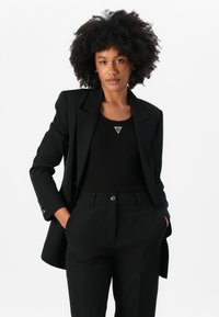 Blazer nero su misura con spalle strutturate, abbinato a una canottiera nera con logo triangolare. Pantaloni neri con tasche.