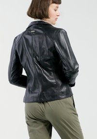 Zwarte leren jack met een aangesloten ontwerp, voorzien van zichtbare stiksels en een puntige kraag. Draagover olijfgroene broek.