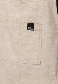 Beige fabric apron with a front pocket. Features stitching details and a small black label with "Jack Wolfskin" in white text.