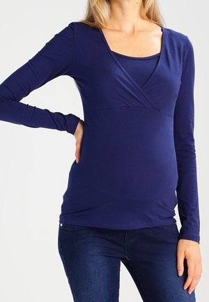 Kobieta ubrana w granatową, długorękawową bluzkę dla kobiet w ciąży z dekoltem w serek oraz ciemnoniebieskie jeansy, z ręką opartą na biodrze.