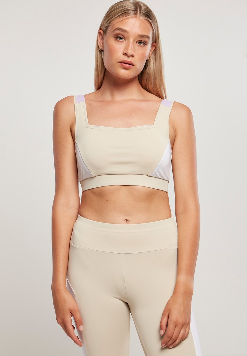 Beige sportovní crop top s čtvercovým výstřihem, kontrastními bílými a levandulovými bočními panely. Hladká textura, přiléhavý střih, nastavitelná ramínka.