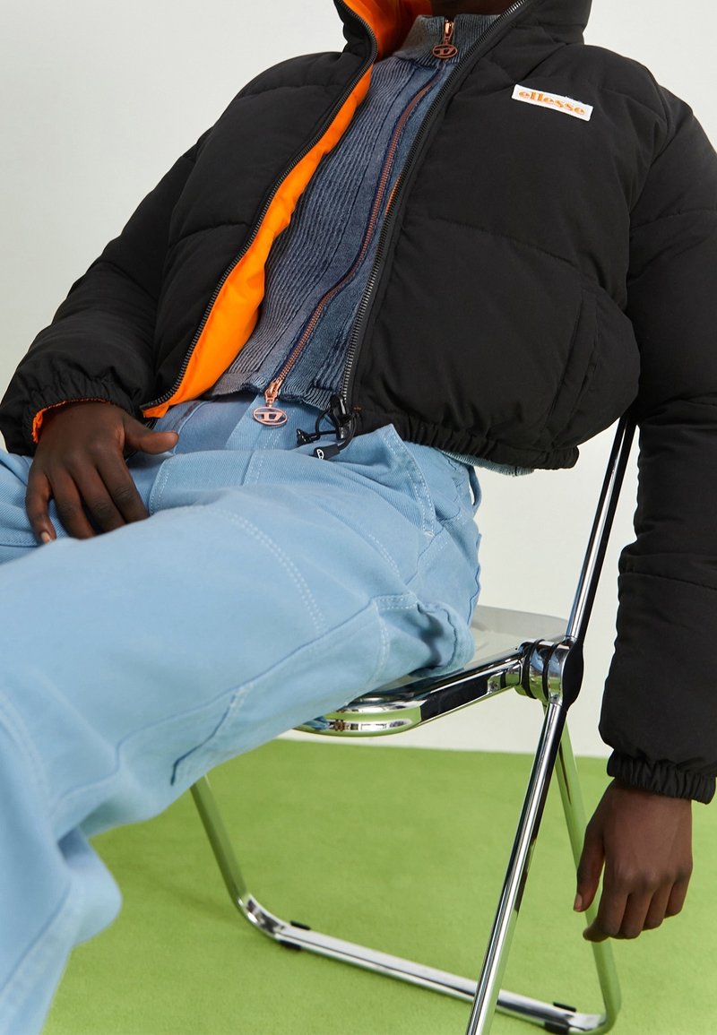 Personne portant une veste zippée noire et orange, un pull côtelé bleu et un pantalon bleu clair, assise sur une chaise en chrome et plastique.