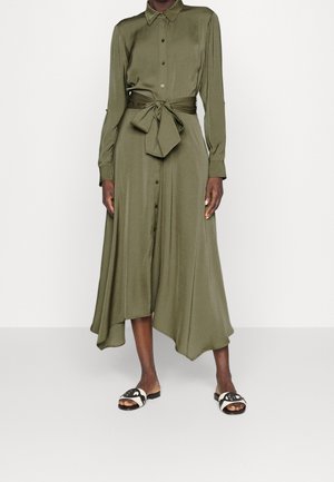 Osoba o ciemnej karnacji ubrana w oliwkowozieloną midi sukienkę na guziki z długimi rękawami i wiązaniem w talii, w połączeniu z czarno-białymi sandałami na płaskim obcasie.