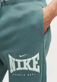 Κοντινό πλάνο σε πετρόλ-πράσινο παντελόνι φόρμας Nike, με χέρι στην τσέπη και κεντημένο λογότυπο "NIKE ATHLETIC DEPT." στο μηρό.