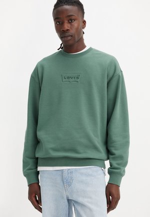Grøn bomuldssweatshirt med rund hals og præget 'Levi's' logo. Har ribbede manchetter og kant. Båret over en hvid skjorte, parret med lyse jeans.