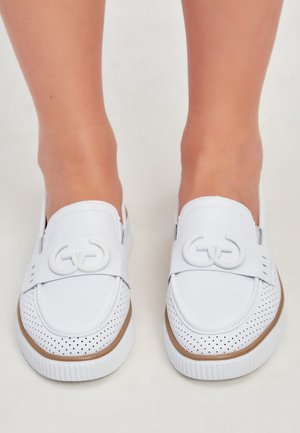 Schuhwerk mit weißen Slip-On-Schuhen mit perforierten Zehenschutzkappen und dekorativem runden Doppelring-Detail oben, getragen an nackten Füßen.