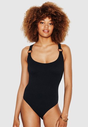 Femme aux cheveux bouclés portant un maillot de bain une pièce noir texturé avec des détails en anneaux sur les bretelles, posant devant un fond uni.