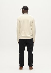 Stickad kreamfärgad tröja med krage, tillsammans med svarta byxor. Modellen bär en svart keps och bruna skor. Minimalistisk design, inga mönster.