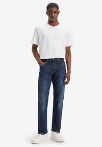 Vaqueros de un azul oscuro con un corte recto, combinados con una camiseta blanca lisa. El modelo adopta una pose relajada, de pie, usando zapatillas blancas.