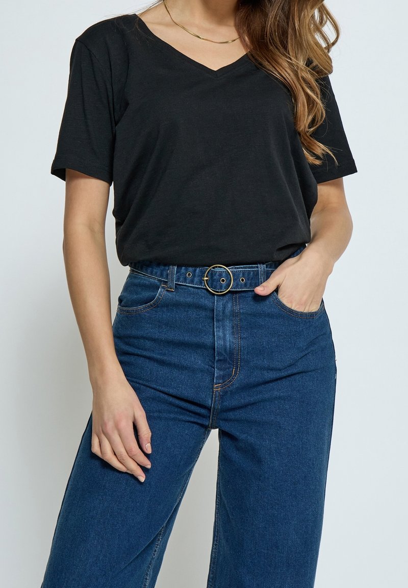 Femme portant un haut noir à manches courtes et encolure en V, rentré dans un jean bleu taille haute avec boucle de ceinture circulaire, main dans la poche.