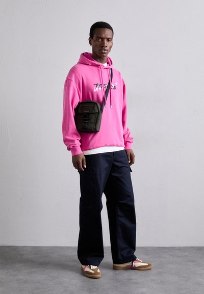 Lys rosa hettegenser med grafisk tekst, mørk marineblå cargo-bukser, grønn crossbody-veske og brune joggesko med rosa detaljer. Glatt stoff og avslappet passform.