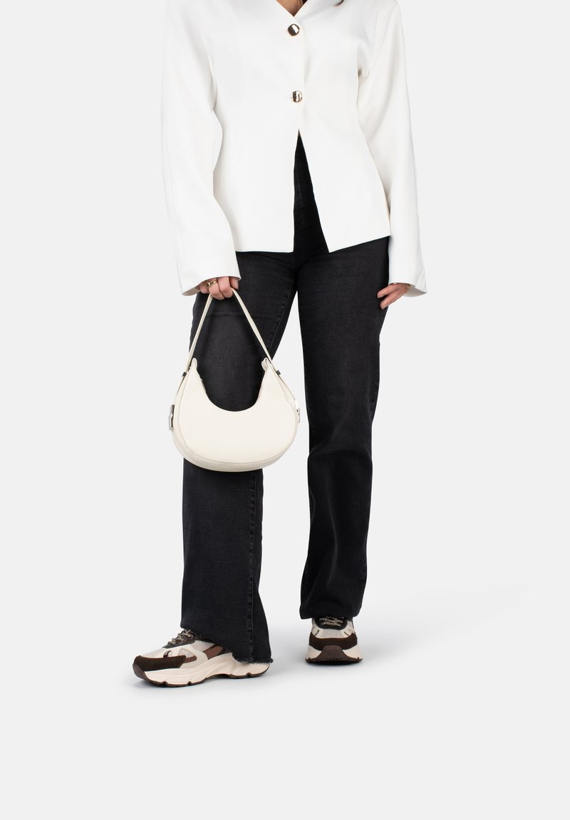 Blazer bianco su misura con bottoni dorati, indossato sopra jeans scuri e sneakers chiare, mentre tiene una piccola borsa hobo bianca dalla forma curva.