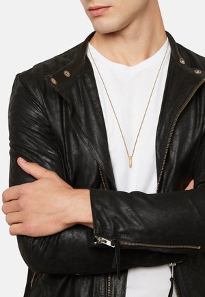 Veste en cuir noir avec fermeture éclair argentée, dotée d'une texture lisse et de détails à pression. T-shirt blanc en dessous et collier avec pendentif doré.