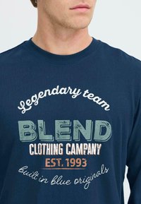 Marineblaues Langarm-T-Shirt mit Text in Weiß, Grün und Orange. Das Design umfasst "Legendäres Team", "BLEND" und weitere Markendetails.