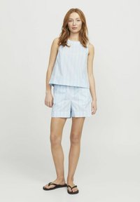 Jauna moteris su raudonais plaukais, vilkinti šviesiai mėlyną dryžuotą be rankovių palaidinę ir atitinkamas šortus, su juodais šlepetėmis, stovinti prieš neutralų foną.