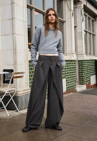 Jasnoszary sweter z mieszanki wełny z okrągłym dekoltem, zestawiony z luźnymi czarnymi spodniami w prążki z plisami i przednimi kieszeniami, noszony z ciemnymi butami.