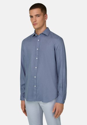 Jeune homme portant une chemise bleue à manches longues boutonnée et un pantalon clair, debout devant un fond uni clair.
