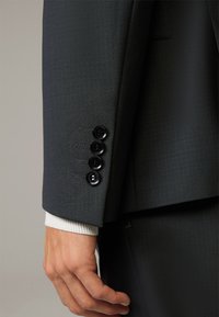 Manica di giacca in un elegante grigio scuro con quattro bottoni neri e una sottile trama a motivo, con un polsino bianco a coste visibile sotto.