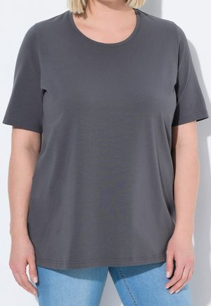 Vrouw met blond haar, gekleed in een effen donkergrijs T-shirt met korte mouwen en lichtblauwe spijkerbroek, staand tegen een witte muur.