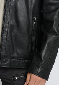 Schwarze Lederjacke mit einer Nahaufnahme eines Reißverschlussfachs und strukturierten Oberflächen, mit einer taillierten Silhouette und geformten Ärmeln.