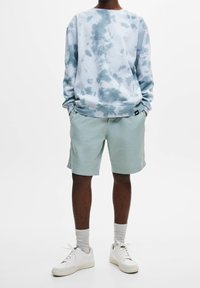 Sweatshirt tie-dye bleu clair avec un col rond, associé à un short vert pâle. Porté avec des baskets blanches et des chaussettes grises à la cheville.