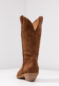 Brune ruskinds cowboystøvler med en spids krave, cirka knæhøje, og en stablet hæl, der viser en glat tekstur og syningsdetaljer.