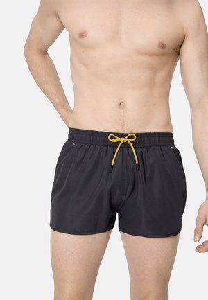 Hombre sin camiseta que lleva pantalones cortos de baño negros con un cordón amarillo, posando frente a un fondo claro y sencillo.